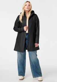 Zwarte waterdichte jas met een capuchon, drukknoopsluiting en zijzakken, gedragen over een lichte top en wijde jeans. Gladde, glanzende textuur.