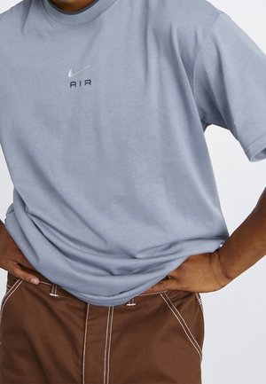 Person iført en lyseblå Nike Air T-shirt og brune bukser med hvide syninger, hænderne hvilende på hofterne.