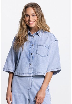 Lachende vrouw met lang, golvend haar, gekleed in een lichtblauw kort spijkerjasje met knopen en bijpassende hooggetailleerde spijkerbroek.