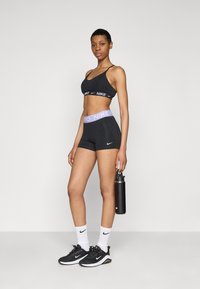 Crni sportski grudnjak sivoj Nike logotip, crne kratke hlače s ljubičastim Nike Pro pojasom, crne tenisice i bijele čarape. Drži crnu bocu vode.