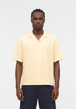 Νεαρός άνδρας με πλεξούδες, που φοράει ανοιχτό κίτρινο κοντομάνικο πουκάμισο με κουμπιά και σκούρα σορτς, στέκεται μπροστά σε απλό φόντο.