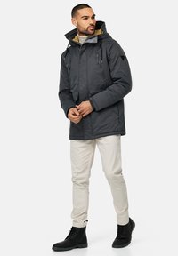 Chaqueta gris aislante con capucha, bolsillos con cremallera y cordones ajustables. Llevada con pantalones claros y botas negras. Tejido texturizado.