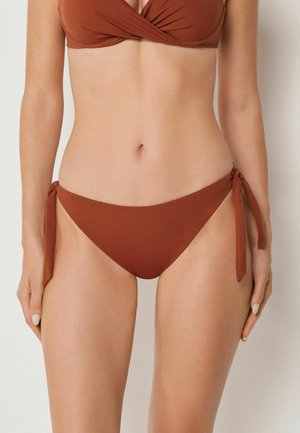 Kvinde iført bikini i rustfarve med sideremme på underdelen og en vrideeffekt foran på overdelen, stående mod en ensfarvet lys baggrund.