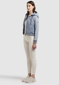 Femme portant un hoodie matelassé bleu clair zippé, des leggings blancs ajustés et des chaussures de course crème, debout devant un fond uni.