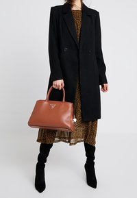 Sac à main en cuir marron avec doubles poignées, forme rectangulaire, logo embossé et finitions en argent, assorti à un manteau noir et une robe à fleurs.