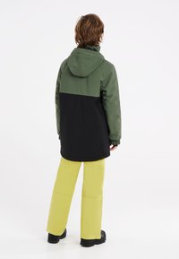 Une veste imperméable bicolore en vert et noir, associée à un pantalon jaune vif. Elle dispose d'une capuche et de poignets élastiques pour le confort.