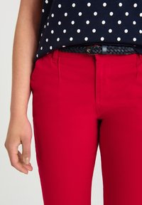 Pantalon rouge ajusté avec une texture lisse, doté de passants pour ceinture et de poches latérales. Associé à un haut bleu marine à pois et une ceinture noire tressée.