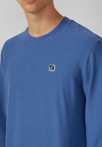 Niebieski sweter dziewiarski o prążkowanej teksturze i okrągłym dekolcie. Posiada małe kwadratowe logo po lewej stronie klatki piersiowej.