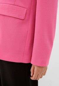 Blazer rose vif en tissu texturé, doté d'une poche à rabat et d'une manche droite. Associé à un pantalon noir.