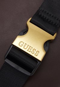 Svart nylonrem med en guldfärgad spänne med den ingraverade "GUESS"-logotypen, mot en brun strukturerad bakgrund.