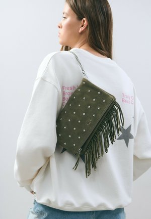 Olive green suede handbag with silver star embellishments, fringe detail, and a chain strap, worn over a beige sweatshirt with printed text.