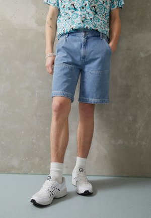 Dongerishorts i en lys blå nyanse, med en klassisk passform, to frontlommer og oppbrettede kanten. Båret med hvite joggesko og sokker.