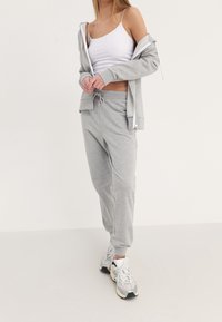 Conjunto de sudadera gris y joggers de tela suave. Presenta un diseño con cremallera, cintura con cordón y puños acanalados. Camiseta blanca debajo.