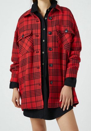 Femme portant une veste à carreaux rouge et noir boutonnée par-dessus une robe noire boutonnée, debout devant un fond blanc.
