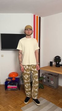 T-shirt oversized bege, calças de padrão camuflado em verde e bege, sapatos pretos. Inclui tatuagens nos braços, boné e características faciais distintas.