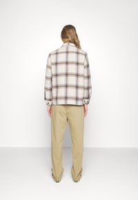 Texturerad krämfärgad jacka med rutmönster, med bruna och blå linjer, bärs med beige byxor. Jackan har rak design och knapppdetaljer.