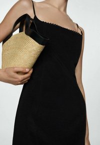 Robe noire à la silhouette ajustée avec des bretelles fines, accompagnée d'un sac en paille doté d'une anse noire et d'une texture tissée.