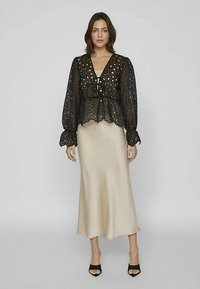 Kobieta ubrana w przezroczystą czarną koronkową bluzkę na beżową sukienkę slip oraz czarne sandały na wysokim obcasie, stojąca na jednolitym szarym tle.