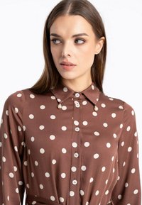 Brązowa bluzka wykonana z gładkiego materiału, z białymi kropkami, kołnierzykiem i zapięciem na guziki z przodu. Wyraźne perłowe akcenty przy kołnierzu.