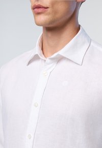 Camicia bianca in lino con colletto button-down, caratterizzata da una sottile texture e un piccolo logo sul petto, con bottoni color perla.