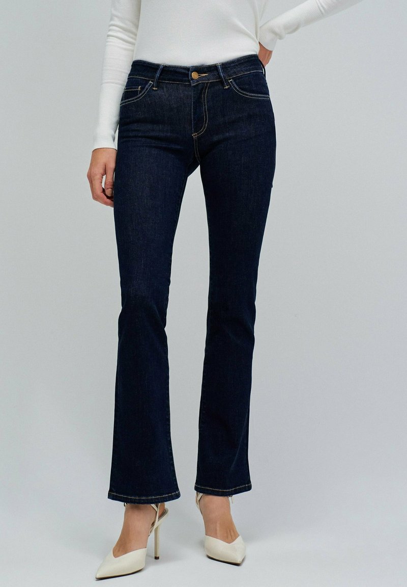 Jeans de mezclilla azul oscuro de corte acampanado con un diseño clásico de cinco bolsillos, herrajes dorados y una cintura a media altura. Textura suave y dobladillo limpio.
