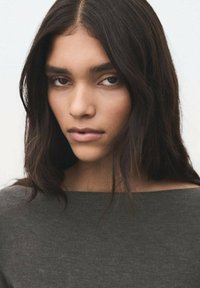 Mørkegrå, ribbet sweater med bådudskæring. Langt, bølget mørkt hår omrammer ansigtet, som har naturlig makeup og subtile highlights.