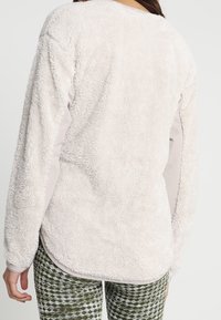 Kobieta ubrana w długi rękaw, kremowy polar i zielone wzorzyste spodnie, widoczna od tyłu na tle jednolitym.