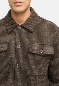Brun corduroy-skjorta med vertikala ränder, med krage, två bröstfickor och grå knappar. Slät textur och skräddarsydd passform.