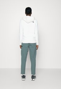 Person som står med ryggen mot kameran iförd vit hoodie, gröna byxor och svarta sneakers mot en enfärgad vit bakgrund.