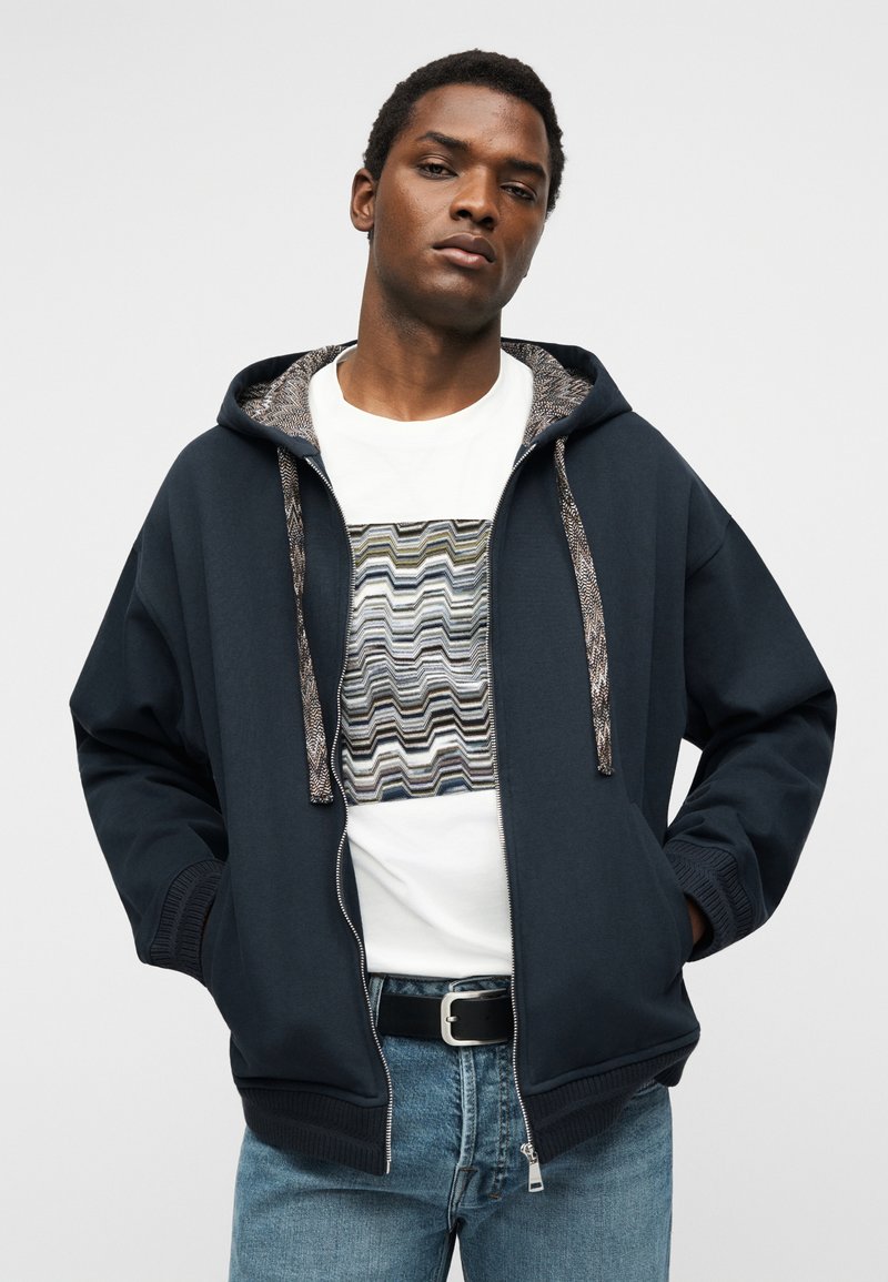 Homme portant un sweat à capuche bleu marine zippé sur un t-shirt blanc imprimé et un jean bleu clair, debout les mains dans les poches sur un fond uni.