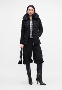 Giacca a vento nera con collo alto, camicia a fantasia, pantaloni cargo neri oversized e stivaletti neri con tacco a spillo al ginocchio. Mini borsa nera.
