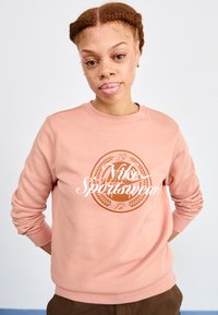 Lys rosa hettegenser laget av mykt materiale, med en sirkulær brun logo som har "Nike Sportswear" og dekorative detaljer foran.