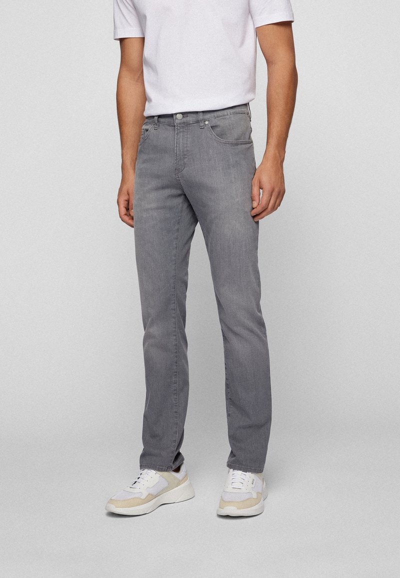 Man med grå slim-fit jeans, vita sneakers och en vit t-shirt med kort ärm, som står framför en enfärgad ljus bakgrund.