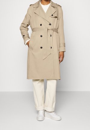 Person trägt einen beigen, doppelreihigen Trenchcoat mit Gürtel, weiße, am Knöchel umgeschlagene Hose und weiße Turnschuhe, steht vor einem schlichten Hintergrund.