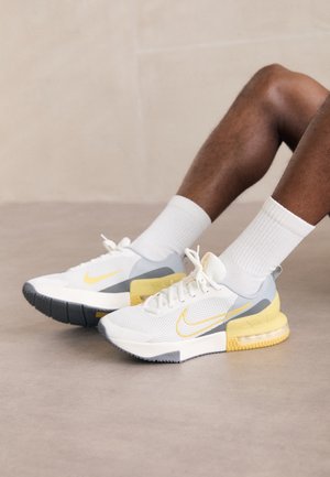Nogi w białych skarpetkach oraz białych, szarych i żółtych butach do biegania Nike, siedzące na gładkiej szarej podłodze.