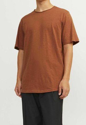 Homme portant un T-shirt uni marron à manches courtes avec un pantalon noir, debout devant un fond blanc.