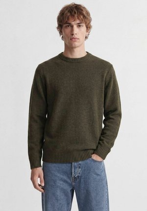Jeune homme aux cheveux bouclés portant un pull en laine vert foncé et un jean bleu, debout avec une main dans la poche devant un fond uni.