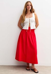 Weißes, mit Spitze verziertes Cropped-Top mit Bindeband an der Vorderseite, kombiniert mit einem langen roten Rock. Braune Sandalen runden das Outfit ab. Einfaches, lässiges Design.