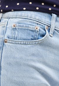 Κοντινό στο τσεπάκι του ανοιχτόμπλε τζιν με μεταλλικά καρφιά και κίτρινη ραφή, σε συνδυασμό με σκουρό μπλε ύφασμα με πουά πάνω από τη μέση.