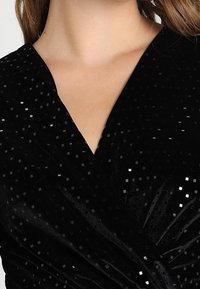 Svart sammetklänning med djup V-ringning, prydd med små glittrande fyrkantiga paljetter som skapar ett texturerat mönster över tyget.