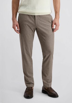 Mann trägt beige schmal geschnittene Hose, braune Ledermokassins und einen hellcremefarbenen, strukturierten Kurzarm-Pullover, steht vor einem weißen Hintergrund.