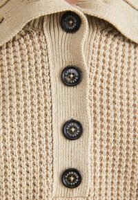 Beige gebreide cardigan met een gestructureerd patroon en vier zwarte knopen met het label "Barbour" op de voorste sluiting.