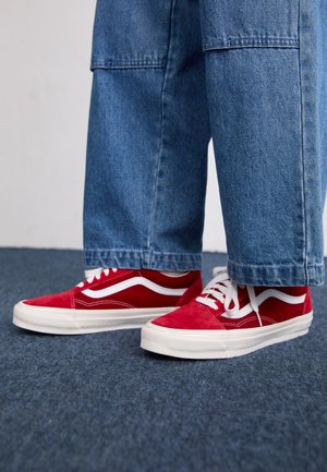 Red suede sneakers with white side stripes, flat rubber soles, and white laces, paired with blue wide-leg denim pants.