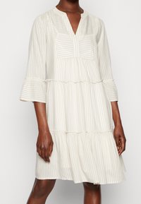 Robe beige clair à rayures verticales blanches, aux manches trois-quarts, avec un ourlet volanté et un col en V, fabriquée en tissu doux et texturé.