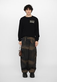 Sort sweater med en tekstureret overflade og "Aries"-logo. Brune cargo-bukser med tie-dye effekt og sorte gummisålesko.