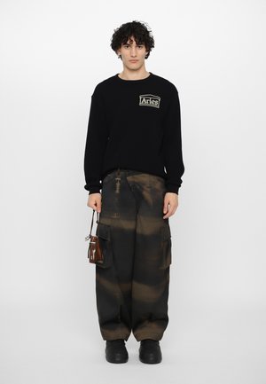 Zwarte trui met een gestructureerde afwerking en "Aries" logo. Bruine, cargostijl broek met een tie-dye effect, en zwarte schoenen met rubberen zolen.