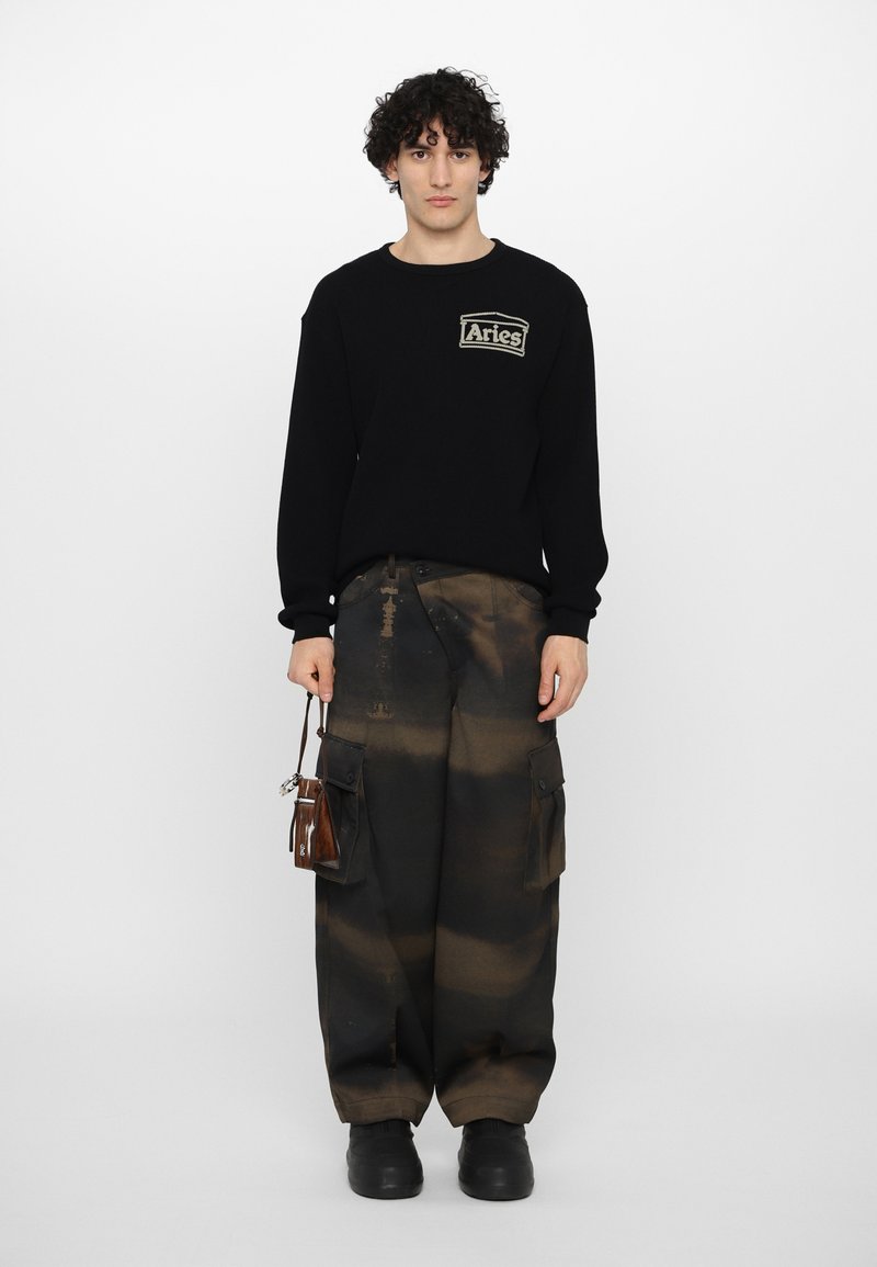 Sort sweater med en tekstureret overflade og "Aries"-logo. Brune cargo-bukser med tie-dye effekt og sorte gummisålesko.