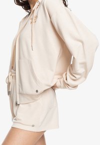 Felpa leggera beige con cappuccio e coulisse, tasca frontale e logo ricamato, abbinata a pantaloni corti coordinati con tasche laterali e dettagli argentati.
