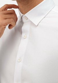 Chemise blanche boutonnée avec col classique, texture lisse et légère brillance. Quatre boutons visibles le long de la patte de boutonnage, design minimaliste.