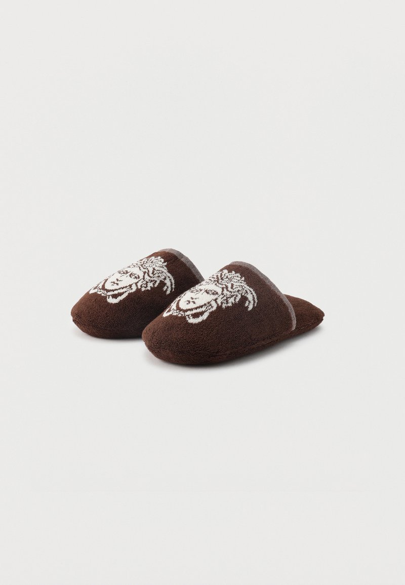 Coppia di pantofole marroni senza lacci con decorazione frontale a forma di volto bianco, realizzate in tessuto morbido e testurizzato.